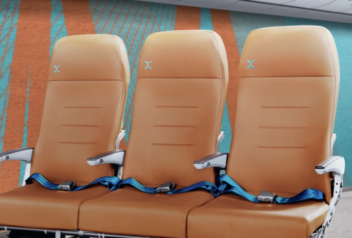 The image shows three adjacent airplane seats with light brown upholstery and blue seatbelts. The seats have armrests and are positioned in front of a colorful wall with orange and teal patterns.