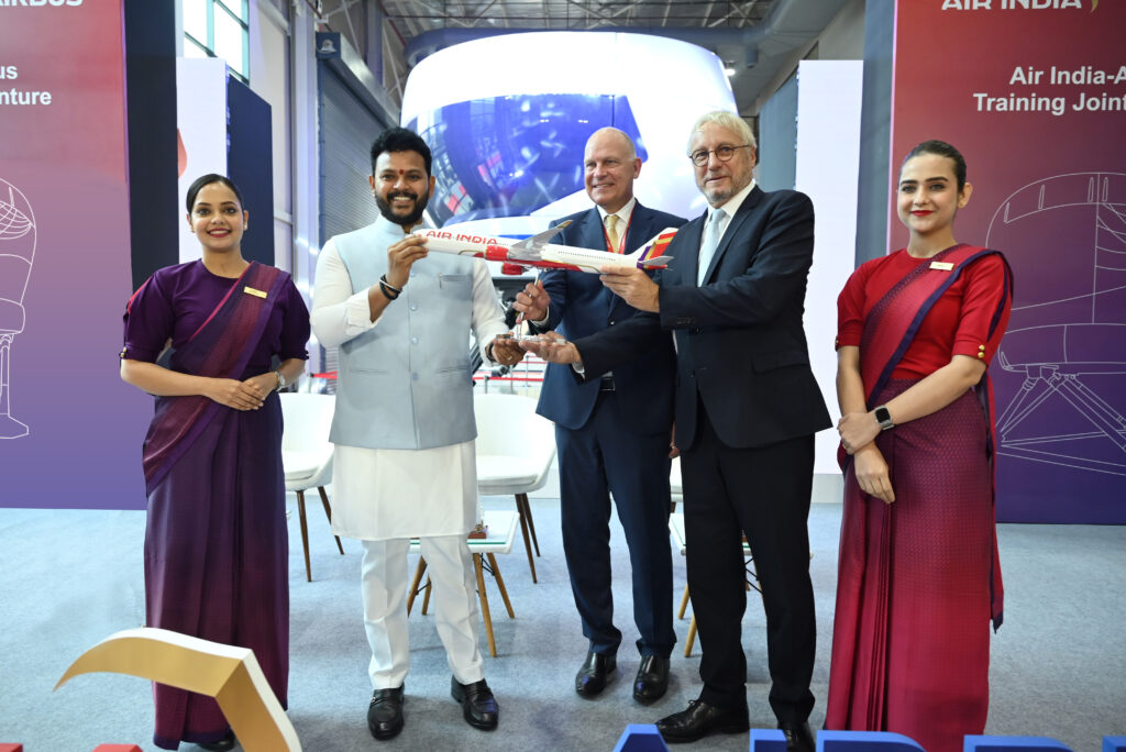 A group of five people is standing indoors, with two women in uniforms on either side and three men in the center. The men are holding a model airplane with "Air India" written on it. The background features banners with "Air India" and "Airbus" logos and text about a training joint venture. The setting appears to be a formal event or announcement.