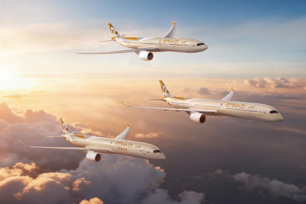Three Etihad Airways airplanes are flying in formation above the clouds during a sunset. The aircraft are painted in the airline's livery, featuring a geometric design on the tail. The sky is a gradient of warm colors, with the sun casting a golden light on the clouds below.