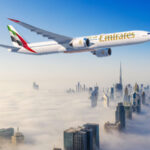 An Emirates airplane is flying over a cityscape with skyscrapers partially covered by clouds. The sky is clear and blue, and the tallest building, resembling the Burj Khalifa, is visible above the clouds.
