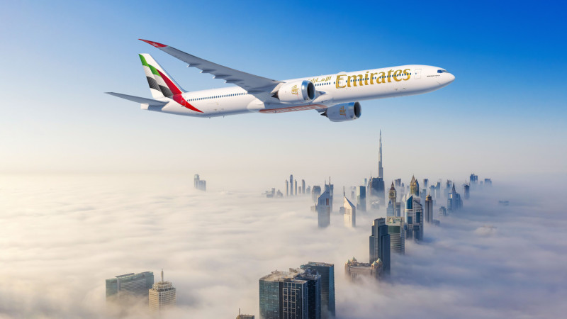 An Emirates airplane is flying over a cityscape with skyscrapers partially covered by clouds. The sky is clear and blue, and the tallest building, resembling the Burj Khalifa, is visible above the clouds.