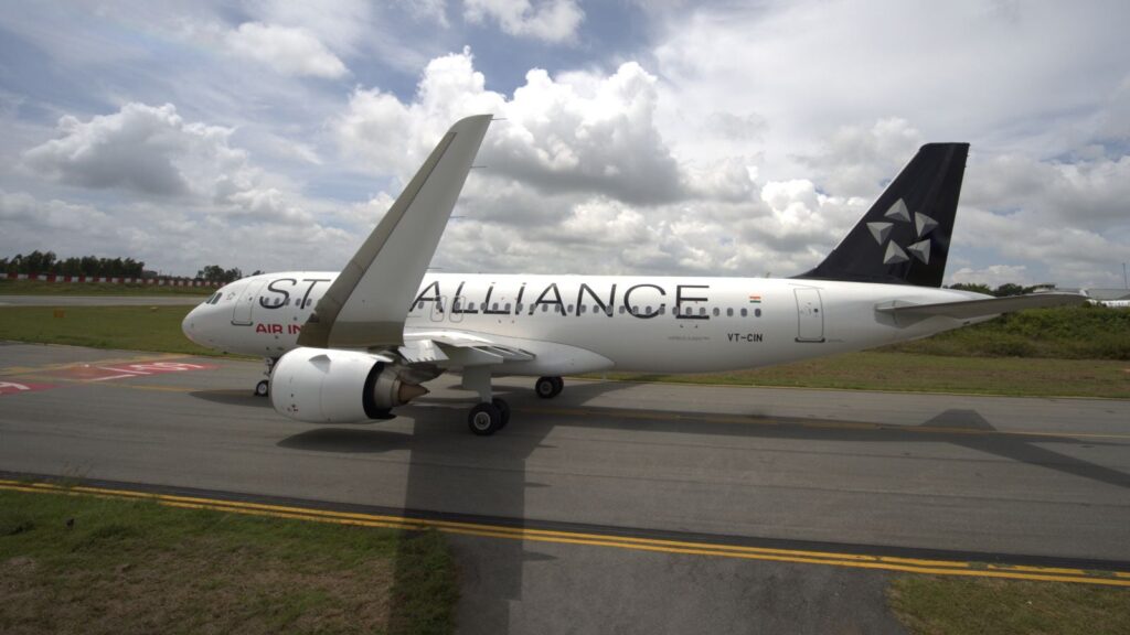 A commercial airplane on a runway, featuring "Star Alliance" branding on its fuselage. The aircraft is white with a dark tail fin displaying a star logo. The sky is partly cloudy, and there is grass alongside the runway.
