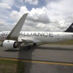 A commercial airplane on a runway, featuring "Star Alliance" branding on its fuselage. The aircraft is white with a dark tail fin displaying a star logo. The sky is partly cloudy, and there is grass alongside the runway.