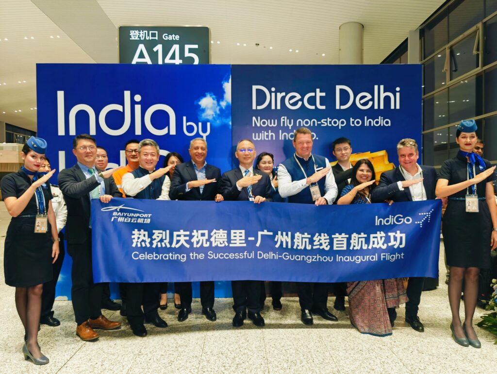 A group of people is standing in an airport terminal in front of a sign that reads "India by IndiGo" and "Direct Delhi." They are holding a banner that says "Celebrating the Successful Delhi-Guangzhou Inaugural Flight" in both English and Chinese. The group includes airline staff and other individuals, some of whom are making a gesture with their arms. The setting is near Gate A145.