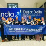 A group of people is standing in an airport terminal in front of a sign that reads "India by IndiGo" and "Direct Delhi." They are holding a banner that says "Celebrating the Successful Delhi-Guangzhou Inaugural Flight" in both English and Chinese. The group includes airline staff and other individuals, some of whom are making a gesture with their arms. The setting is near Gate A145.