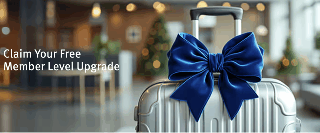 A silver suitcase with a large blue bow is in the foreground, set in a blurred hotel lobby. The text on the left reads, "Claim Your Free Member Level Upgrade."