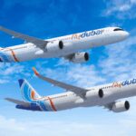 The image shows two commercial airplanes in flight against a blue sky with scattered clouds. The planes are white with blue and orange accents, and they have the "flydubai" logo on their bodies and tails. The aircraft are flying in the same direction, with one slightly above the other.