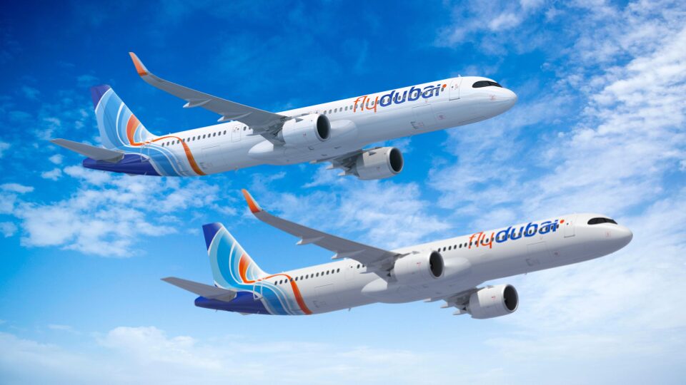 The image shows two commercial airplanes in flight against a blue sky with scattered clouds. The planes are white with blue and orange accents, and they have the "flydubai" logo on their bodies and tails. The aircraft are flying in the same direction, with one slightly above the other.