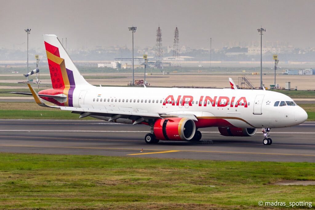 The image shows an Air India airplane on a runway at an airport. The aircraft is white with red and gold accents, and the Air India logo is visible on the fuselage. The tail features a colorful design with red, purple, and gold patterns. The background includes airport infrastructure and a hazy cityscape.