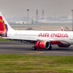 The image shows an Air India airplane on a runway at an airport. The aircraft is white with red and gold accents, and the Air India logo is visible on the fuselage. The tail features a colorful design with red, purple, and gold patterns. The background includes airport infrastructure and a hazy cityscape.