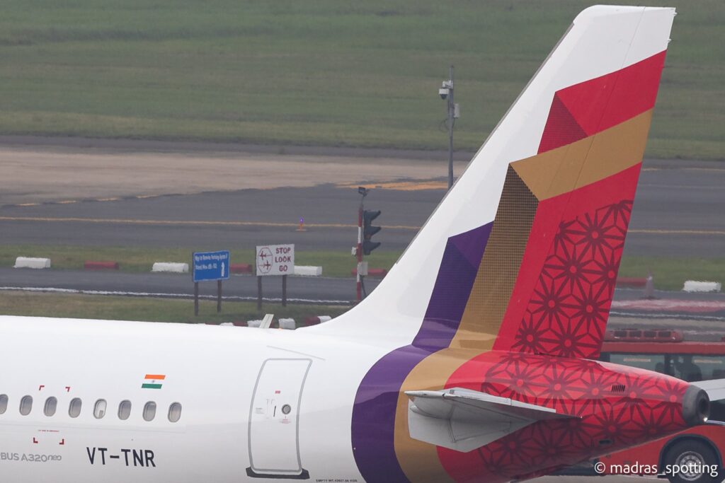 The image shows the tail section of an Airbus A320neo aircraft on a runway. The tail features a colorful geometric design in red, purple, and gold. The Indian flag is visible on the fuselage, along with the registration number "VT-TNR." In the background, there are signs and a grassy area near the runway.