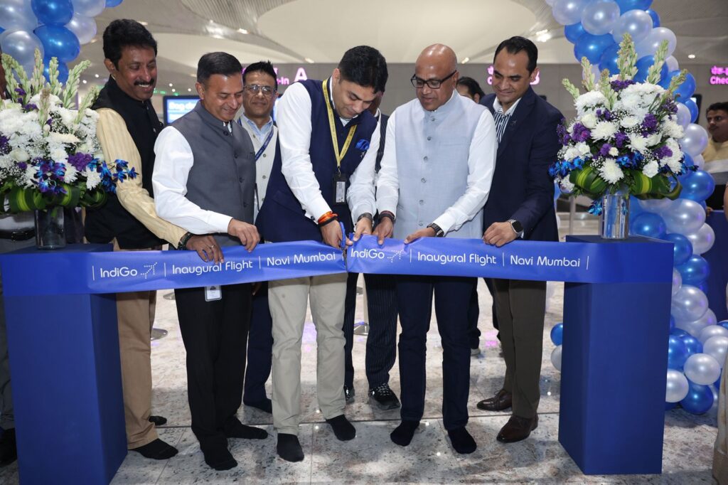 A group of men is participating in a ribbon-cutting ceremony for an inaugural flight at an airport. They are cutting a blue ribbon that reads "IndiGo Inaugural Flight Navi Mumbai." The scene is decorated with blue and white balloons and floral arrangements. The men are dressed in formal attire, and the setting appears to be inside an airport terminal.
