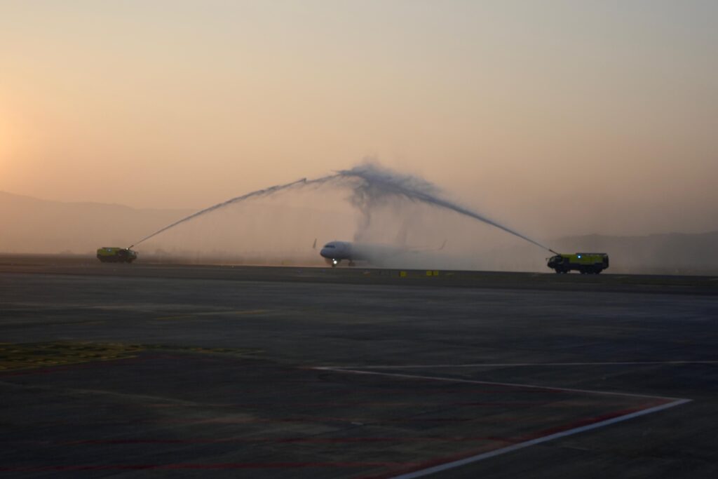 An airplane is on a runway being greeted with a water salute from two fire trucks, one on each side. The scene is set during sunset or sunrise, with a warm, hazy sky in the background.