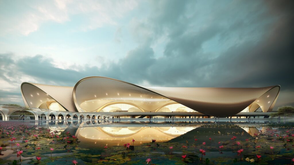 The image shows a modern architectural structure with a sweeping, curved roof design. The building is reflected in a body of water in the foreground, which is adorned with pink water lilies. The sky above is partly cloudy, creating a dramatic backdrop. The structure features large, arched openings and appears to be illuminated from within.