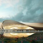 The image shows a modern architectural structure with a sweeping, curved roof design. The building is reflected in a body of water in the foreground, which is adorned with pink water lilies. The sky above is partly cloudy, adding a dramatic backdrop to the scene. The structure features large, illuminated arches and appears to be a public or cultural building.