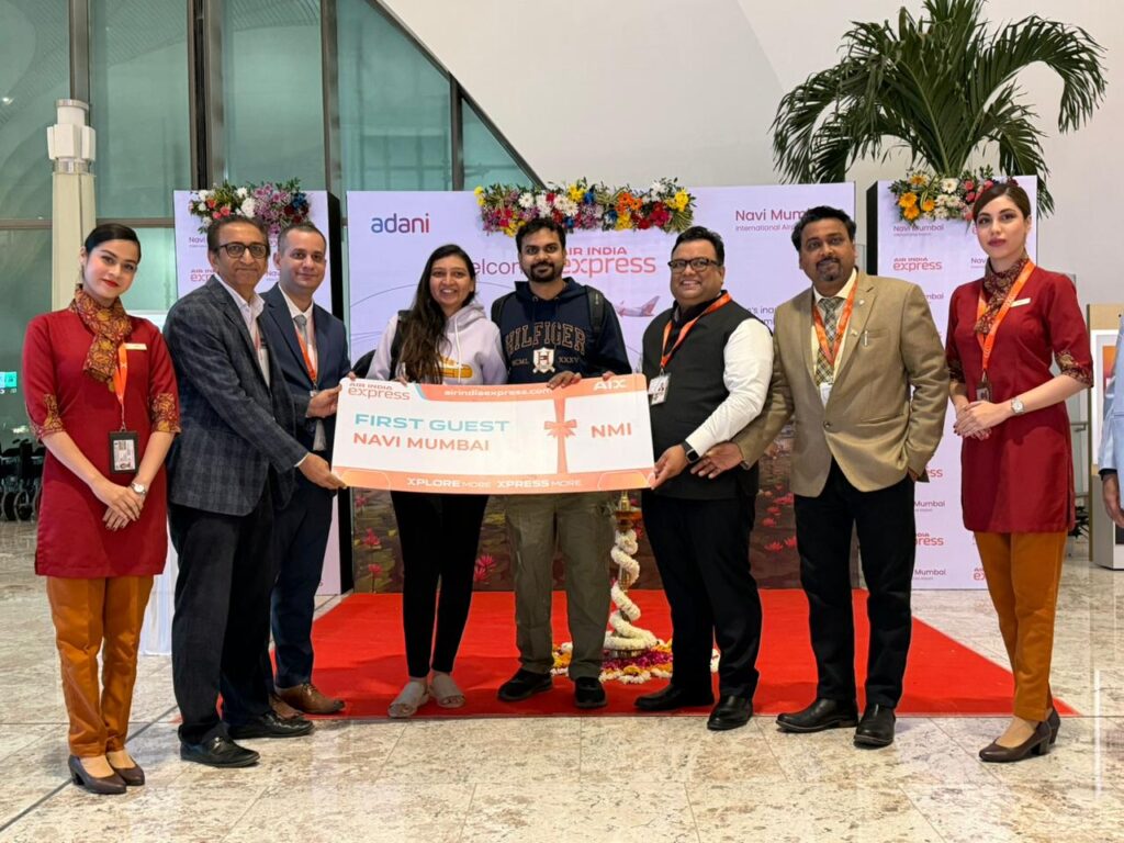 A group of people is standing on a red carpet at an airport, holding a large sign that reads "First Guest Navi Mumbai." They are smiling and appear to be part of a welcoming ceremony. The background features banners with the Air India Express logo and floral decorations. Two women in red uniforms are standing on either side of the group.