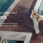 A couple is walking hand in hand on a wooden boardwalk surrounded by water, with tropical plants nearby. The woman is wearing a sun hat and a long dress, while the man is in a white shirt and shorts. Text on the image promotes a 40% bonus points offer and mentions the Sheraton Grand Mirage Resort, Gold Coast.