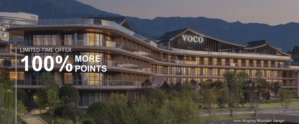The image shows a modern, multi-story hotel building with large windows and balconies, set against a backdrop of mountains. The hotel has the name "VOCO" displayed prominently on the top. In the foreground, there are landscaped gardens with trees and shrubs. Text on the left side of the image reads "LIMITED-TIME OFFER 100% MORE POINTS." The bottom right corner has the text "voco Wugong Mountain Jiangxi."