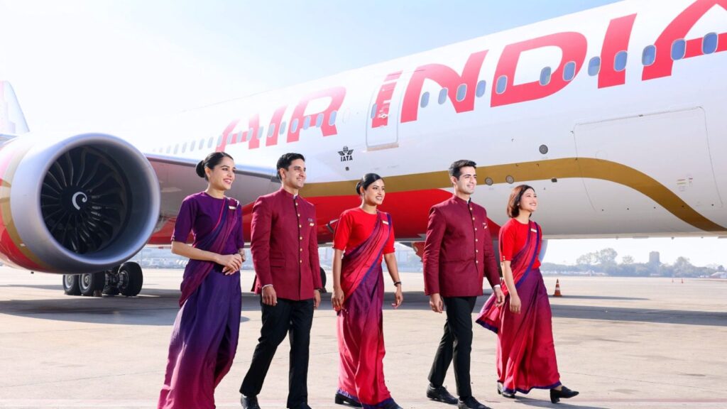 The image shows five airline crew members walking on the tarmac in front of an Air India airplane. They are wearing coordinated uniforms in shades of red and purple. The airplane's engine and part of the fuselage with the "Air India" logo are visible in the background. The crew members are smiling and appear to be in a professional setting.