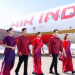 The image shows five airline crew members walking on the tarmac in front of an Air India airplane. They are wearing coordinated uniforms in shades of red and purple. The airplane's engine and part of the fuselage with the "Air India" logo are visible in the background. The crew members are smiling and appear to be in a professional setting.