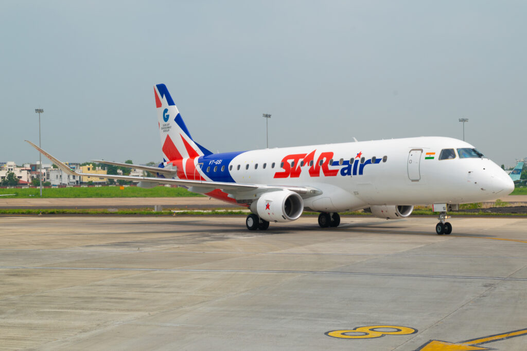 The image shows a Star Air passenger airplane on the tarmac at an airport. The aircraft is white with red and blue accents, featuring the Star Air logo prominently on the fuselage. The tail has a design with red, white, and blue colors, and the Indian flag is visible near the cockpit. The background includes a grassy area and some buildings.