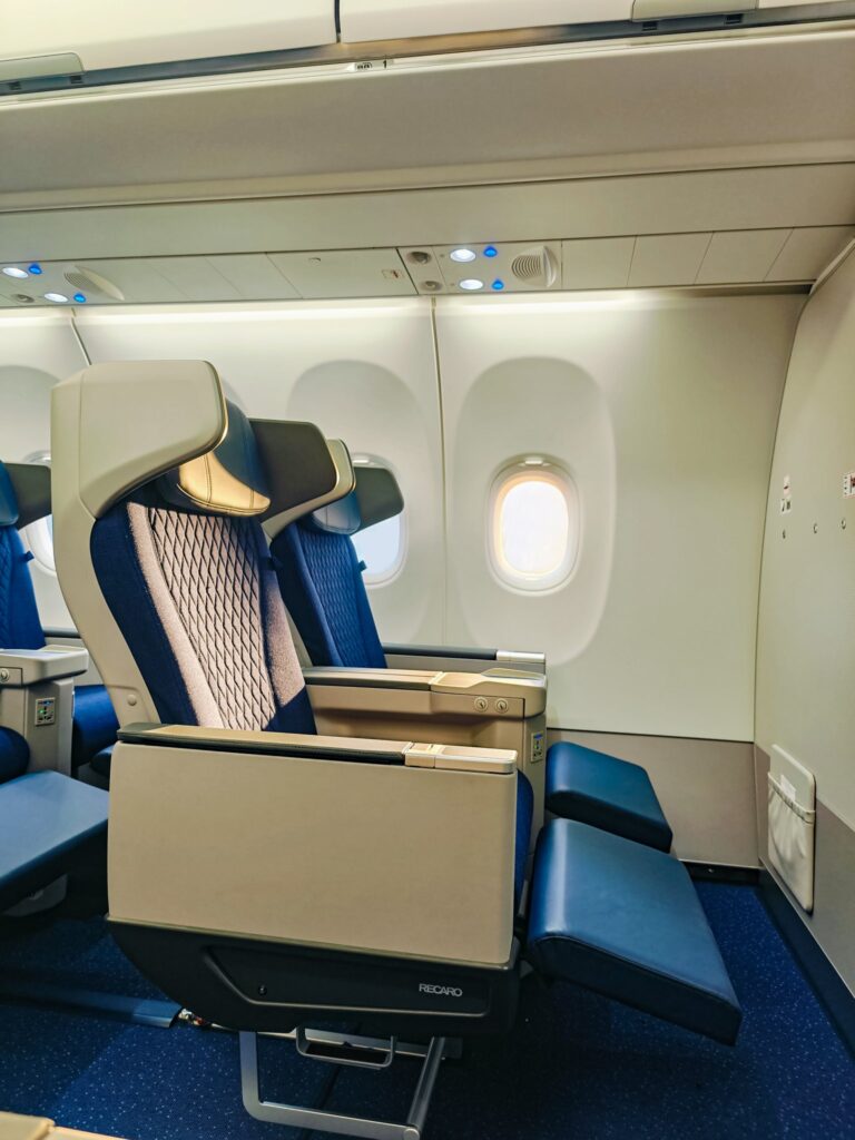 The image shows the interior of an airplane cabin, focusing on a row of business class seats. The seats are upholstered in blue fabric with a patterned design and have adjustable leg rests. Overhead compartments and individual reading lights are visible above the seats. The cabin has a clean and modern appearance.