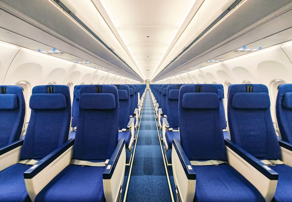 The image shows the interior of an empty airplane cabin. There are rows of blue seats on either side of a central aisle. The overhead compartments are closed, and the lighting is bright, giving a clean and orderly appearance.