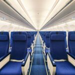 The image shows the interior of an empty airplane cabin. There are rows of blue seats on either side of a central aisle. The overhead compartments are closed, and the lighting is bright, giving a clean and orderly appearance.