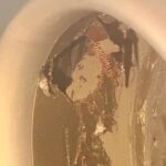 The image shows a close-up of a damaged or corroded metallic surface inside a circular object, possibly a pipe or a container. The surface has visible scratches, peeling, and areas of discoloration, revealing different layers of material. The lighting casts shadows, highlighting the texture and irregularities on the surface.