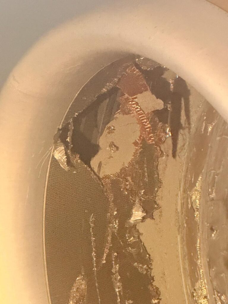 The image shows a close-up of a damaged or corroded metallic surface inside a circular object, possibly a pipe or a container. The surface has visible scratches, peeling, and areas of discoloration, revealing different layers of material. The lighting casts shadows, highlighting the texture and irregularities on the surface.