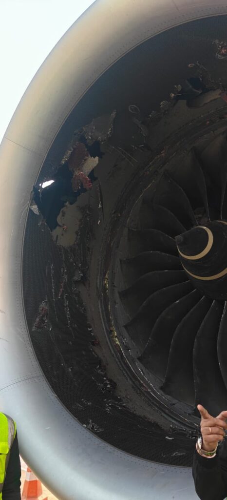The image shows a close-up of a damaged aircraft engine. The engine's fan blades and inner components are visible, with noticeable damage and debris around the edges. A person in a high-visibility vest is partially visible on the left side, and another person's hand is pointing at the engine on the right side.
