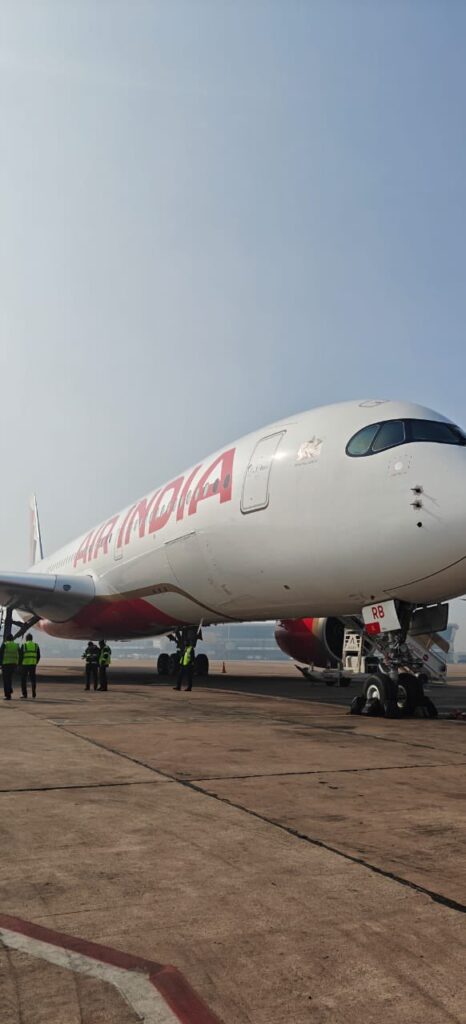 The image shows a large Air India airplane parked on an airport tarmac. Several people wearing high-visibility vests are standing near the aircraft. The sky is clear and blue.