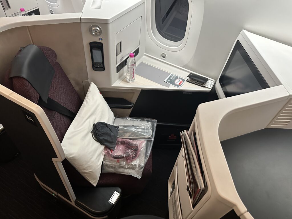 The image shows a first-class airplane seat setup. The seat is upholstered in dark fabric and has a seatbelt. A white pillow and a plastic-wrapped blanket are placed on the seat. A small black pouch is also on the seat. The area includes a personal entertainment screen, a bottle of water, and a control panel. There is a window next to the seat, and various compartments and storage spaces are visible.