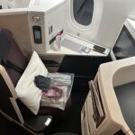 The image shows a first-class airplane seat setup. The seat is upholstered in dark fabric and has a seatbelt. A white pillow and a plastic-wrapped blanket are placed on the seat. A small black pouch is also on the seat. The area includes a personal entertainment screen, a bottle of water, and a control panel. There is a window next to the seat, and various compartments and storage spaces are visible.