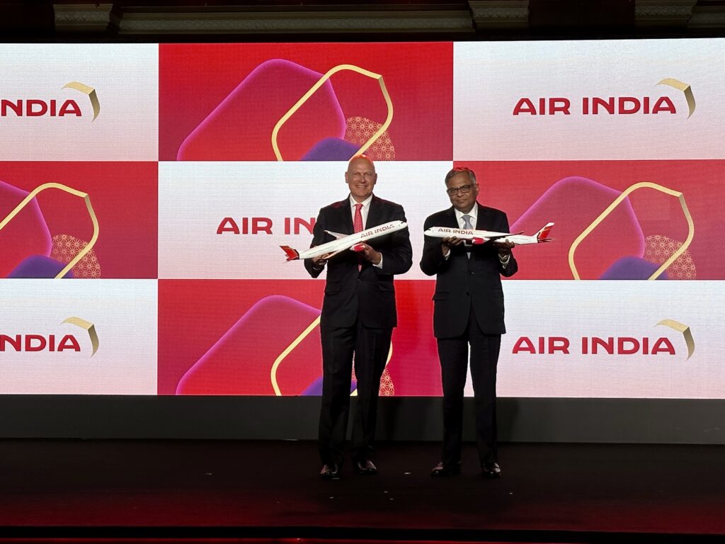 Two men in suits are standing on a stage holding model airplanes. The background displays the Air India logo multiple times on a red and white backdrop.