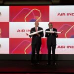 Two men in suits are standing on a stage holding model airplanes. The background displays the Air India logo multiple times on a red and white backdrop.