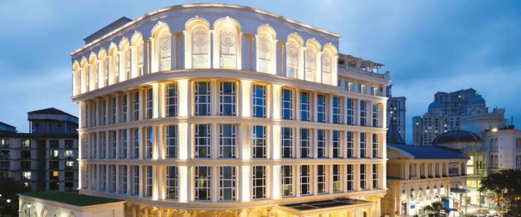 The image shows a large, elegant building with multiple stories, featuring a classical architectural style. The facade is illuminated with warm lighting, highlighting its arched windows and decorative elements. The building is situated in an urban area with other structures visible in the background under a dusky sky.