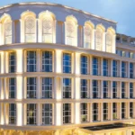 The image shows a large, elegant building with multiple stories, featuring a classical architectural style. The facade is illuminated with warm lighting, highlighting its arched windows and decorative elements. The building is situated in an urban area with other structures visible in the background under a dusky sky.