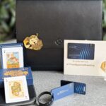 The image shows a collection of items displayed on a table. There is a black leather bag with a gold keychain attached, a deck of playing cards with some cards visible, a black zippered pouch, and a cardholder displaying a "Maharaja Club" membership card. Next to these is a brochure with the text "#ThatSpecialFeeling" and a logo. There is also a commemorative coin in a clear case, featuring an airplane design and the year "1932." The background is blurred with greenery.