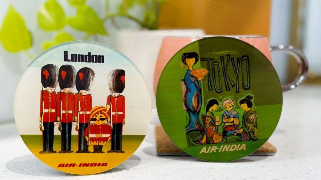 The image shows two round coasters with vintage Air India designs. The left coaster features cartoon illustrations of British guards in red uniforms and bearskin hats, with the word "London" above them. The right coaster depicts a stylized scene of people in traditional Japanese attire, with the word "Tokyo" above. Both coasters have "Air-India" written at the bottom. In the background, there are blurred plants and a mug.
