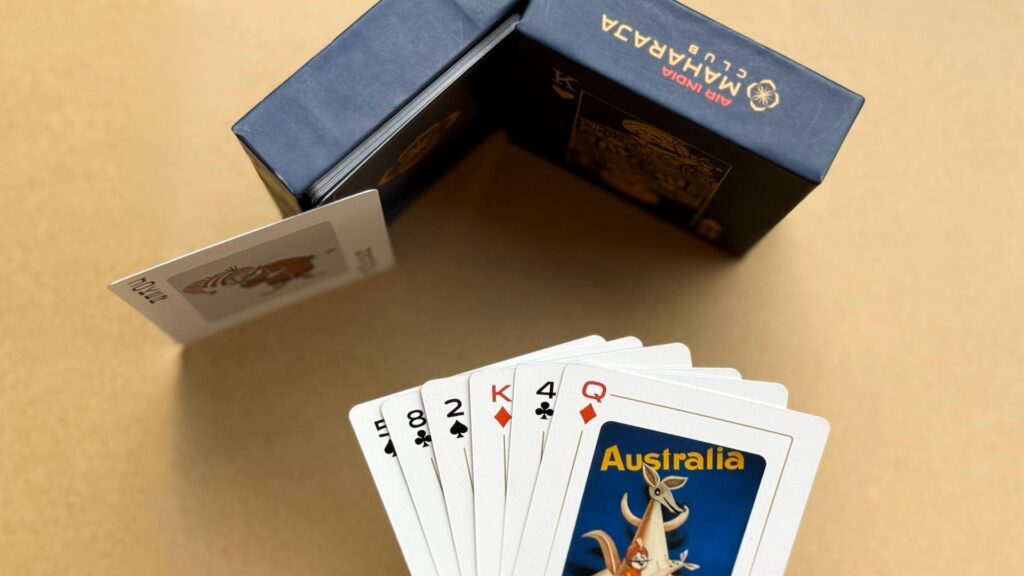 The image shows a set of playing cards fanned out on a surface. The visible cards include the 5, 8, 2, King, and Queen of clubs, and the 4 of diamonds. One card features an illustration with the word "Australia" and a kangaroo. In the background, there is a blue box with the text "Air India Maharaja Club" partially visible.