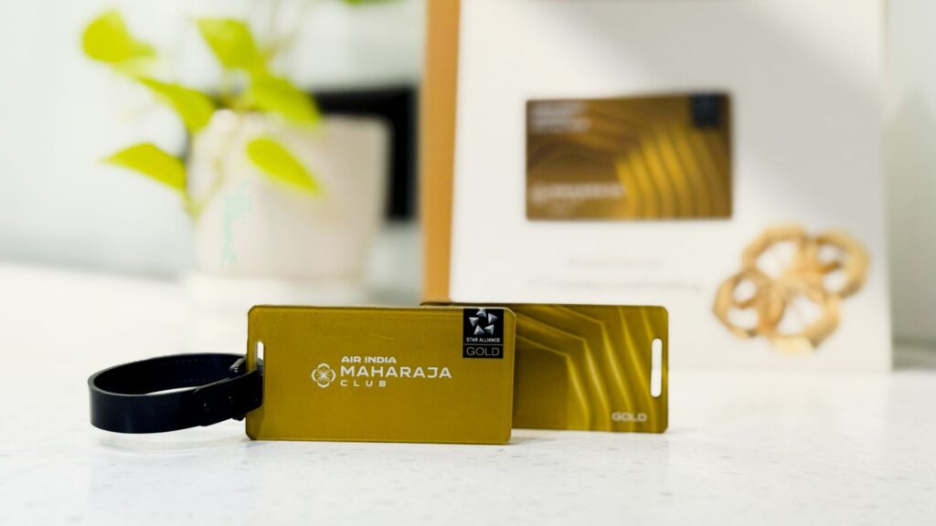 The image shows a gold-colored Air India Maharaja Club luggage tag with a black strap. The tag features the Star Alliance Gold logo. In the background, there is a blurred box with a similar design and a plant with green leaves.