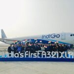 A group of people is standing in front of an IndiGo airplane on an airport tarmac. They are holding Indian flags and posing for a photo. In front of them is a sign that reads "India's first A321XLR." The airplane is white with blue accents and the IndiGo logo on the side. The scene is set on a clear day.