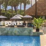 The image shows a luxurious outdoor pool area at the Kimpton Surfcomber Hotel. The pool is surrounded by lounge chairs and umbrellas, with lush greenery and palm trees in the background. A thatched-roof structure is visible, adding to the tropical ambiance. Text on the image reads "Limited-Time Offer 100% More Points."