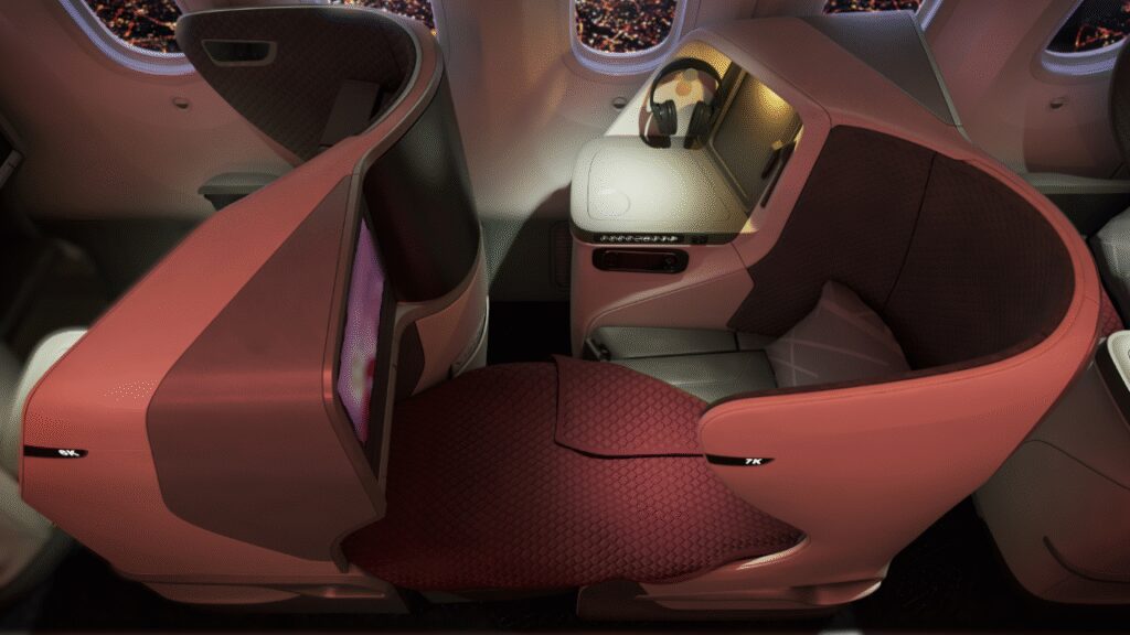 This image shows a luxurious airplane business class seat. The seat is spacious and features a curved privacy shell with a quilted design. It includes a large screen, a side table with a pair of headphones, and a control panel. The seat is upholstered in a combination of red and gray materials, and there is a window with a view of city lights outside.