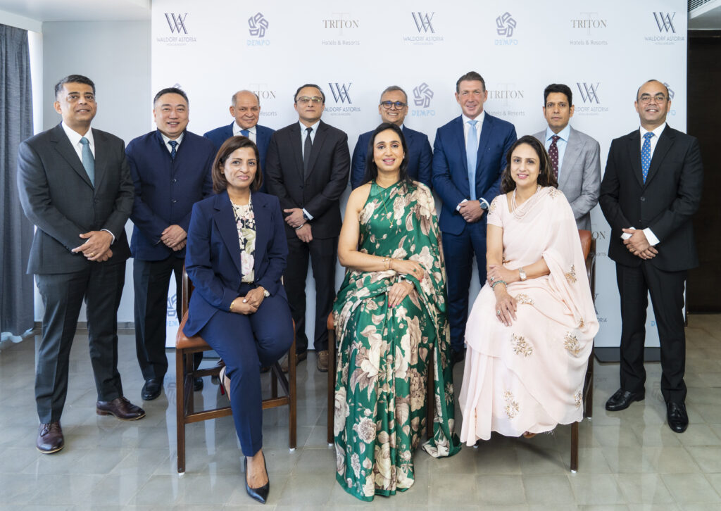 A group of eleven people, consisting of eight men and three women, are posing for a formal photograph. The men are wearing suits, while the women are dressed in elegant attire, with one in a floral green dress and another in a light pink dress. They are standing and sitting in two rows against a backdrop featuring logos of Waldorf Astoria, Dempo, and Triton Hotels & Resorts.