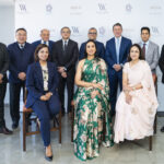 A group of eleven people, consisting of eight men and three women, are posing for a formal photograph. The men are wearing suits, while the women are dressed in elegant attire, with one in a floral green dress and another in a light pink dress. They are standing and sitting in two rows against a backdrop featuring logos of Waldorf Astoria, Dempo, and Triton Hotels & Resorts.