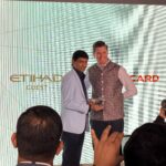 Two men are standing together on a stage, smiling and holding a glass award. One is wearing a light blue blazer, and the other is in a patterned vest. Behind them, logos for "Etihad Guest" and "Mastercard" are visible. Several people in the foreground are taking photos.