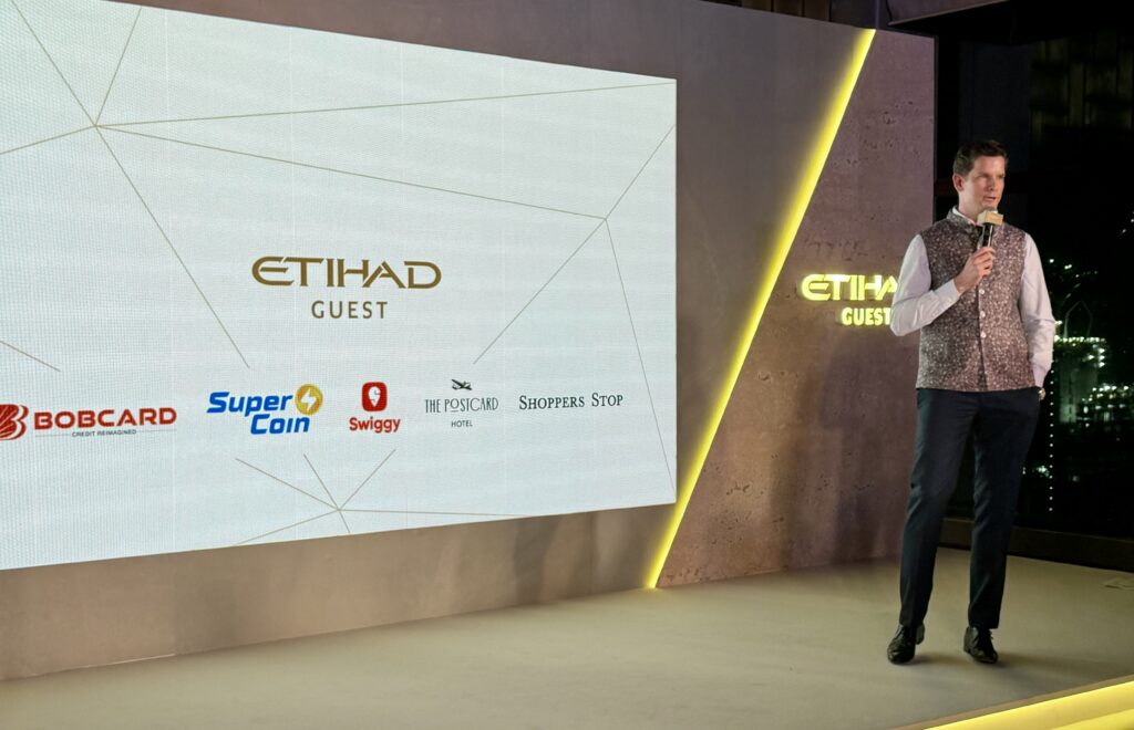 A person is standing on a stage holding a microphone, next to a large screen displaying the "Etihad Guest" logo. Below the logo are several other logos, including Bobcard, Super Coin, Swiggy, The Postcard Hotel, and Shoppers Stop. The stage has a modern design with a yellow-lit edge.