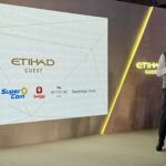 A person is standing on a stage holding a microphone, next to a large screen displaying the "Etihad Guest" logo. Below the logo are several other logos, including Bobcard, Super Coin, Swiggy, The Postcard Hotel, and Shoppers Stop. The stage has a modern design with a yellow-lit edge.